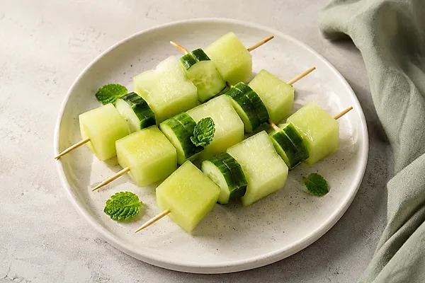 Kleine Spieße mit Melonenwürfeln und Gurkenscheiben auf einem hellen Teller.